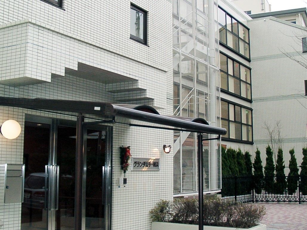 白いタイルの外壁で、窓が並んでいる大規模な建物である。白い外階段が見えている。きれいに立ち並んだ植樹がある。