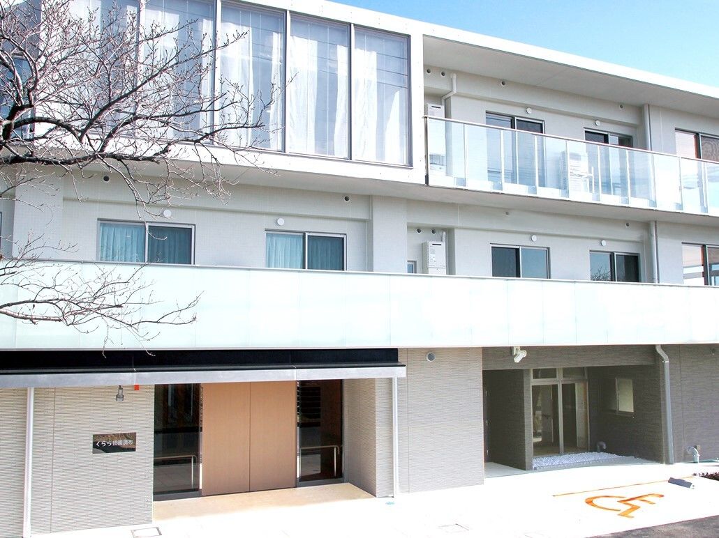 3階建てで白い外観の施設の前には大きな木が１本植えられている。入口には屋根があり、日よけや雨よけになる。