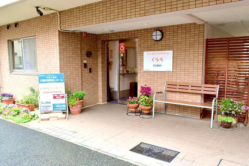 鉄筋コンクリート３階建ての施設入口には木製ベンチが置かれ、上部の壁には施設名称のプレートが設置されている。