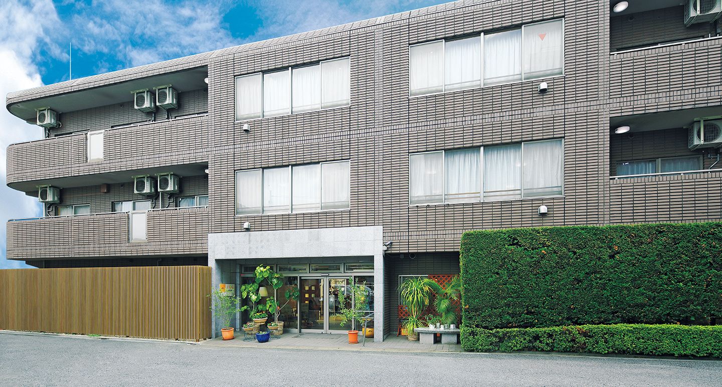 濃い茶色の壁の建物である。入り口部分は白い壁になっており、バリアフリー構造である。外には緑がたくさん植えられている。