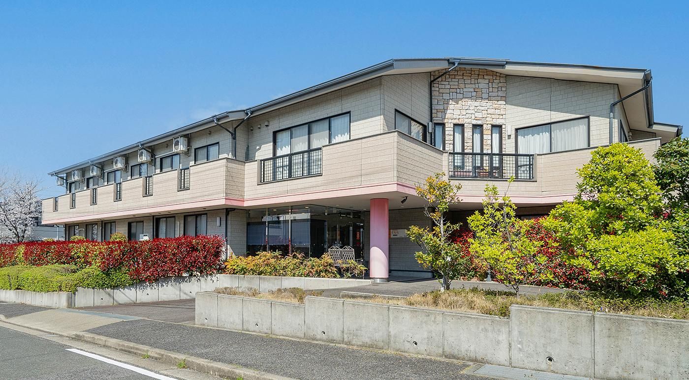 灰色の落ち着いた色合いにピンクの支柱が特徴的な、２階建ての施設である。入口前方には鮮やかな色の花や草木が生い茂っている。