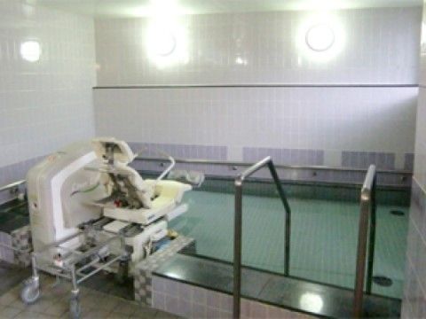 床と壁がタイル貼りの浴室に、大きな浴槽がある。周囲に手すりがあり、座ったまま入浴する介助器具が付いている。