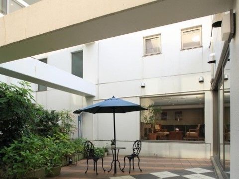 施設敷地内に設けられたテラスである。青いパラソルが付いたテーブルと１人掛けの椅子が２脚置かれている。