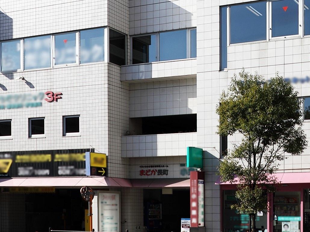 赤い文字で書かれた施設名が目印の白い建物。併設されたクリニックの一階部分は駐車場になっており、建物中央の外壁には植物がある。