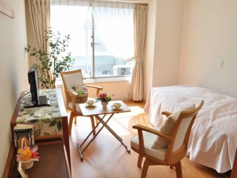 窓があり、壁と床がベージュで明るい部屋は、一方の壁際にベッドがある。中央に椅子２脚とテーブルがあり、反対側の壁には整理棚が２つ並んでいる。