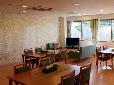 黒っぽい色の座面の椅子がセットされたダイニングテーブルが、通路に余裕を持っていくつも並べられている。部屋の角にテレビとソファが置かれている。
