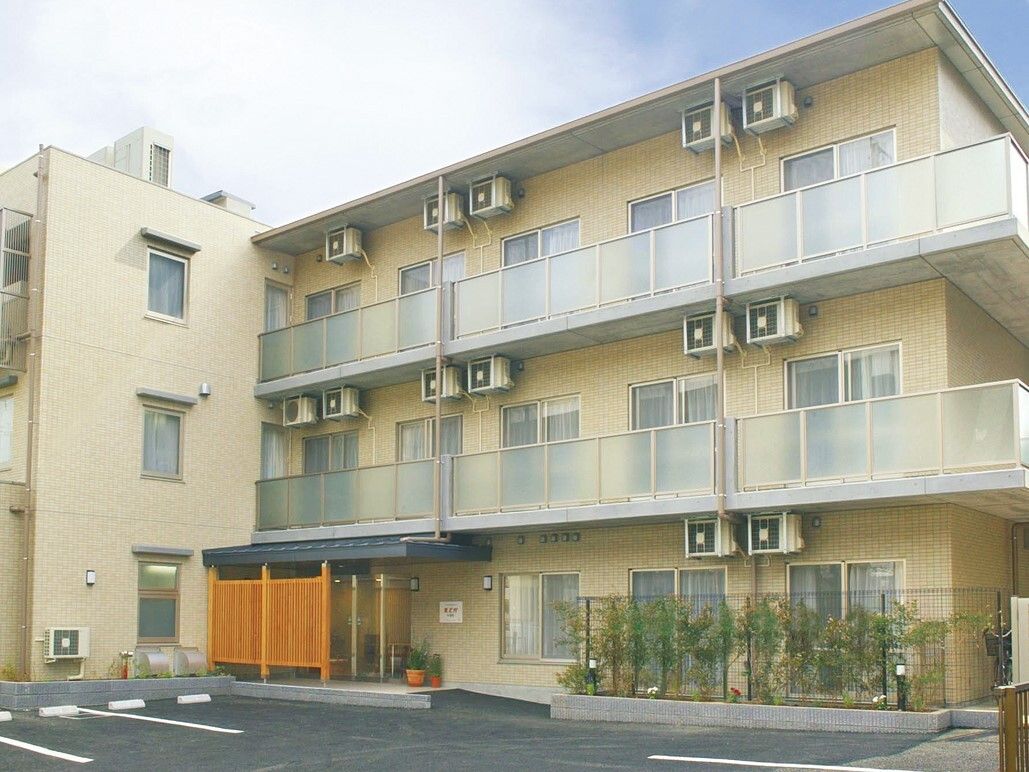 ３階建ての建物で、１階の周りにはフェンスや植物がある。敷地内は舗装されており、駐車場が整備されている。