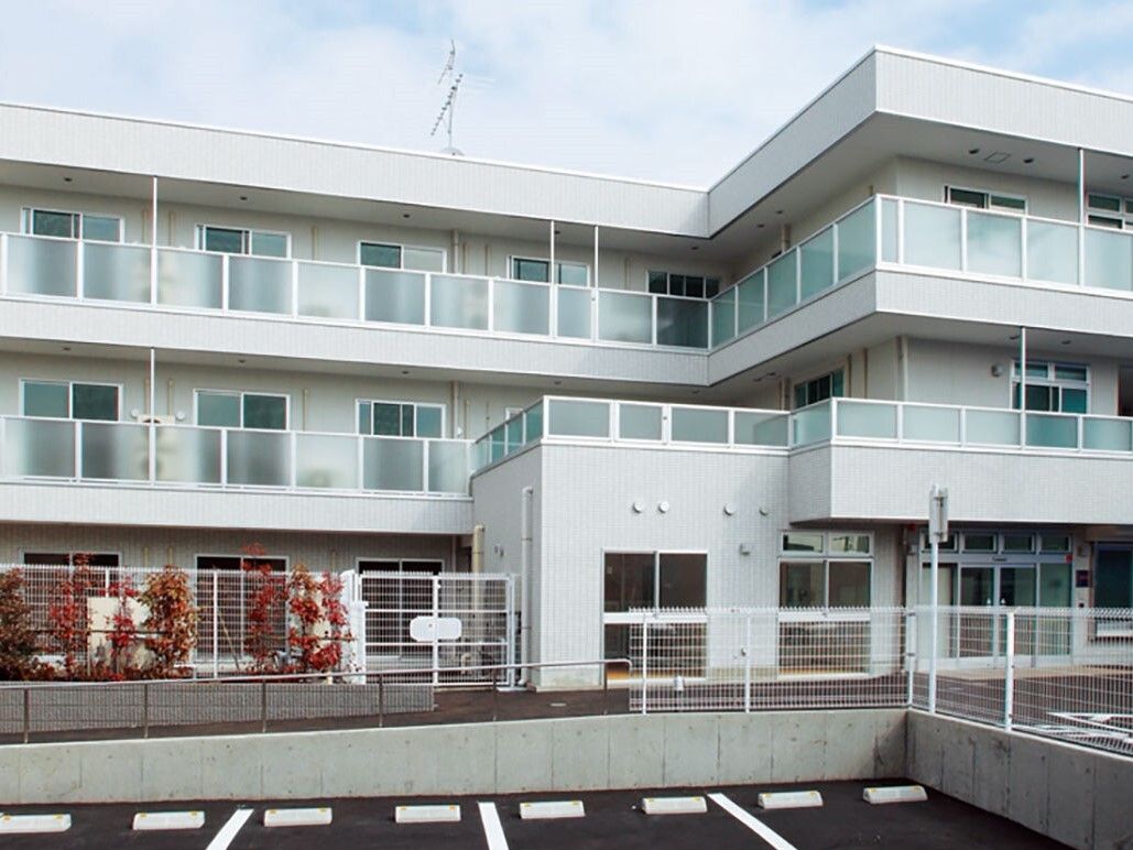 ３階建ての建物は白を基調としている。玄関の両脇にはコニファーと花の寄せ植えがある。緑のパラソルがあるガーデンチェアもある。
