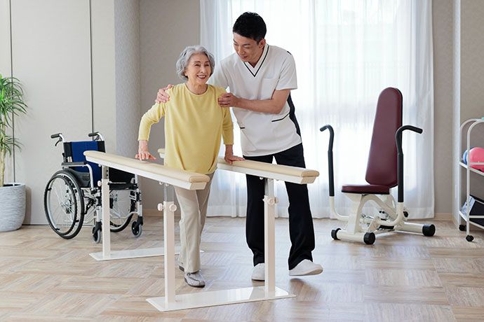 カルテを手に高齢者に話しかける白衣の女性と床にかがむ男性スタッフ