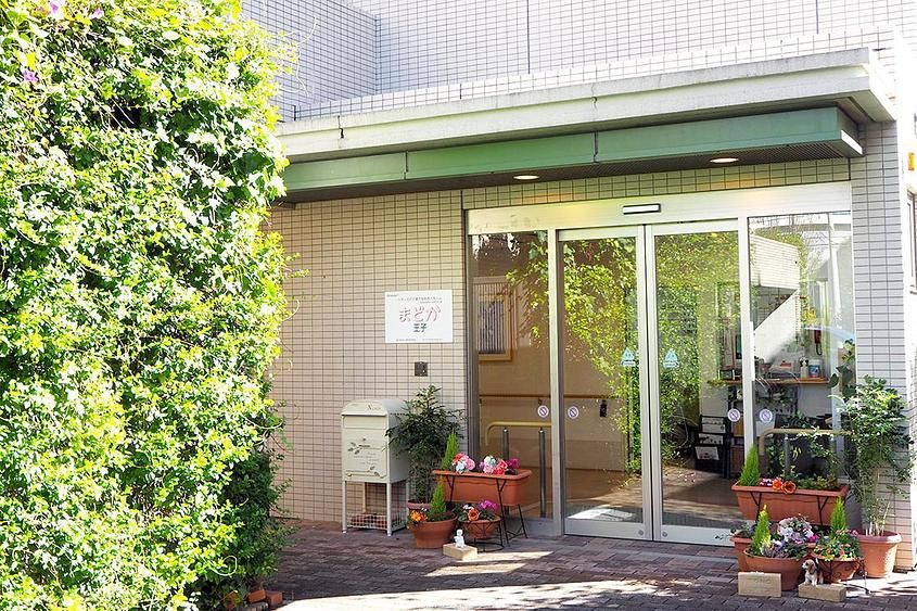 玄関はガラスの自動扉で、入り口横に施設名が表示されている。玄関マットが敷かれている。壁に沿って、植物が這っている。
