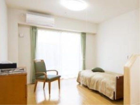 大きめのベッドが備え付けられた明るいフローリングの居室である。天井近くの壁にはエアコンが取り付けられ、大きな窓からは日の光が差し込む。