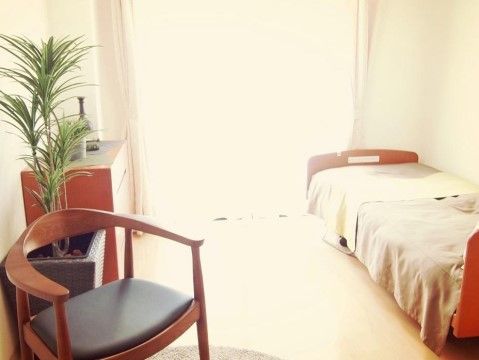 居室の壁際にベッドが置かれている。大きな観葉植物の鉢植えが飾られている。大きな窓から外光が差し込んでいる。