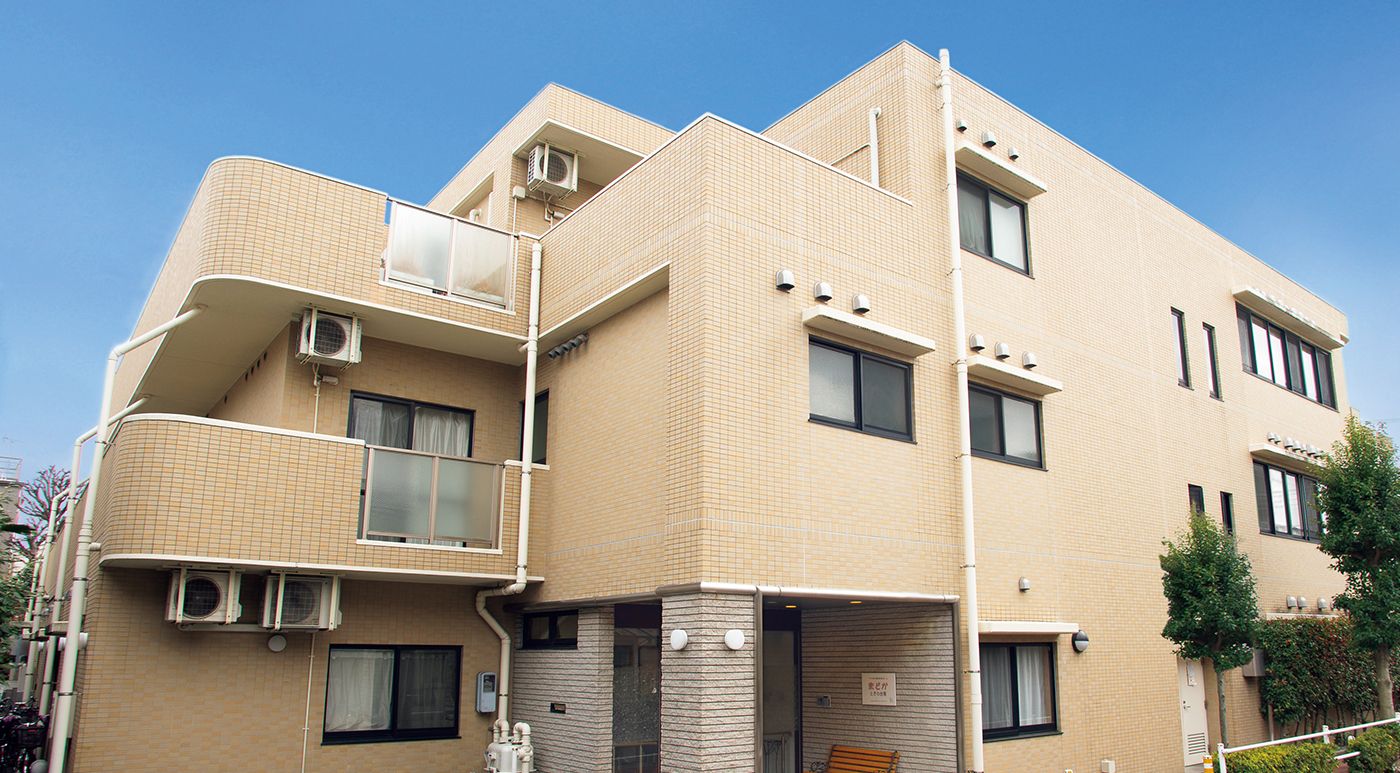施設は３階建てで薄いベージュの建物である。玄関は屋根がついている。施設前に駐車スペースが用意されている。