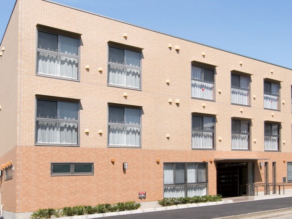 ３階建てで優しい色使いの外観。施設入口周辺には手すりが取り付けられている。目の前の道路が綺麗に舗装されている。