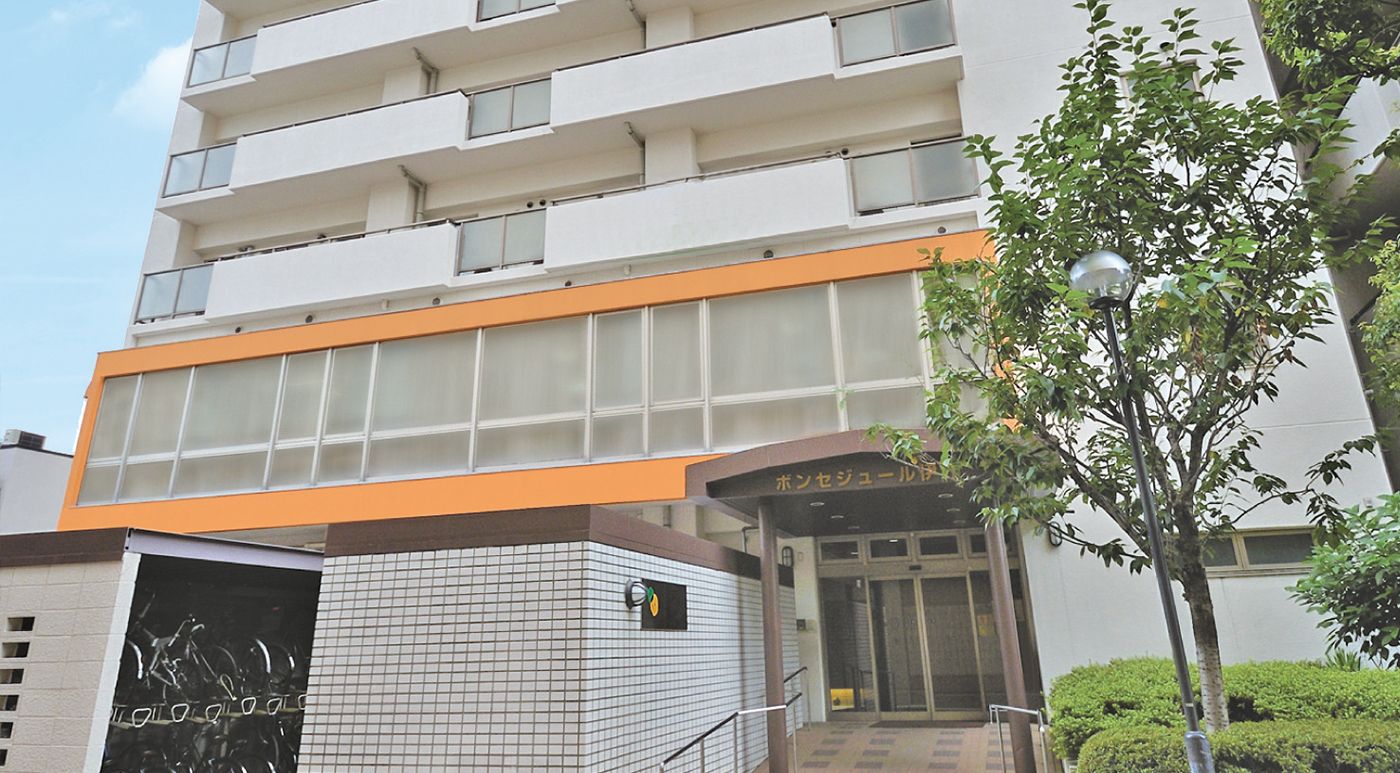 白い外壁のマンションのような縦に大きい建物である。周囲には街灯が設置され、木が植えられている。２階部分は窓がたくさんある。