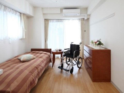 部屋の中心に黒い車椅子が置いてある。その横にベッドがあり、赤のしま模様のシーツの上に白いクッションが載っている。