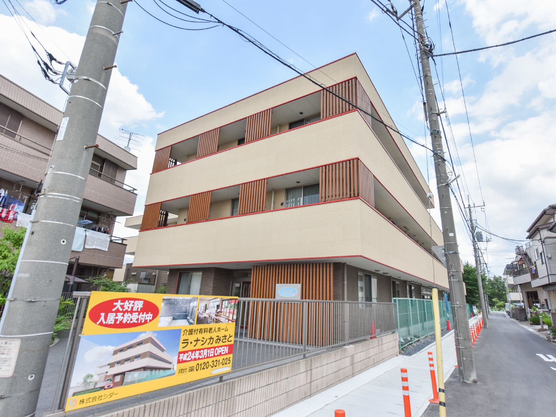 3階建ての建物の出っ張った壁の部分はクリーム色で塗られている。建物は柵で囲まれており、そのすぐ横は道路が通っている。