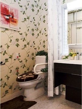 植物をモチーフにした壁紙のトイレ。トイレの横には可動式の手すりが付けられ、隣にある洗面台との間にはカーテンが取り付けられている。