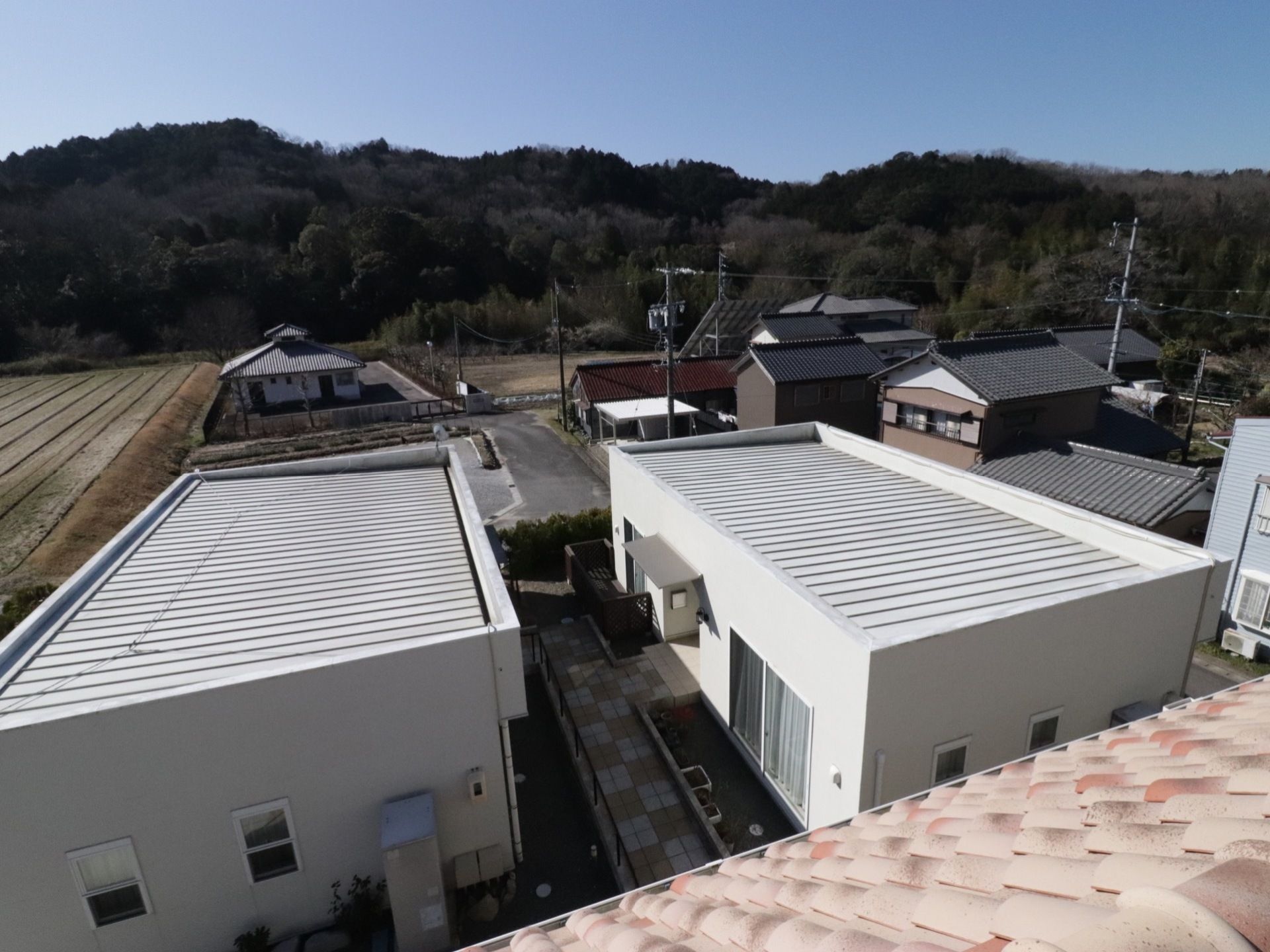 青空に山々が広がる、のどかな景色である。手前に白い漆喰の壁の棟が２棟見える。奥には民家が建っている。