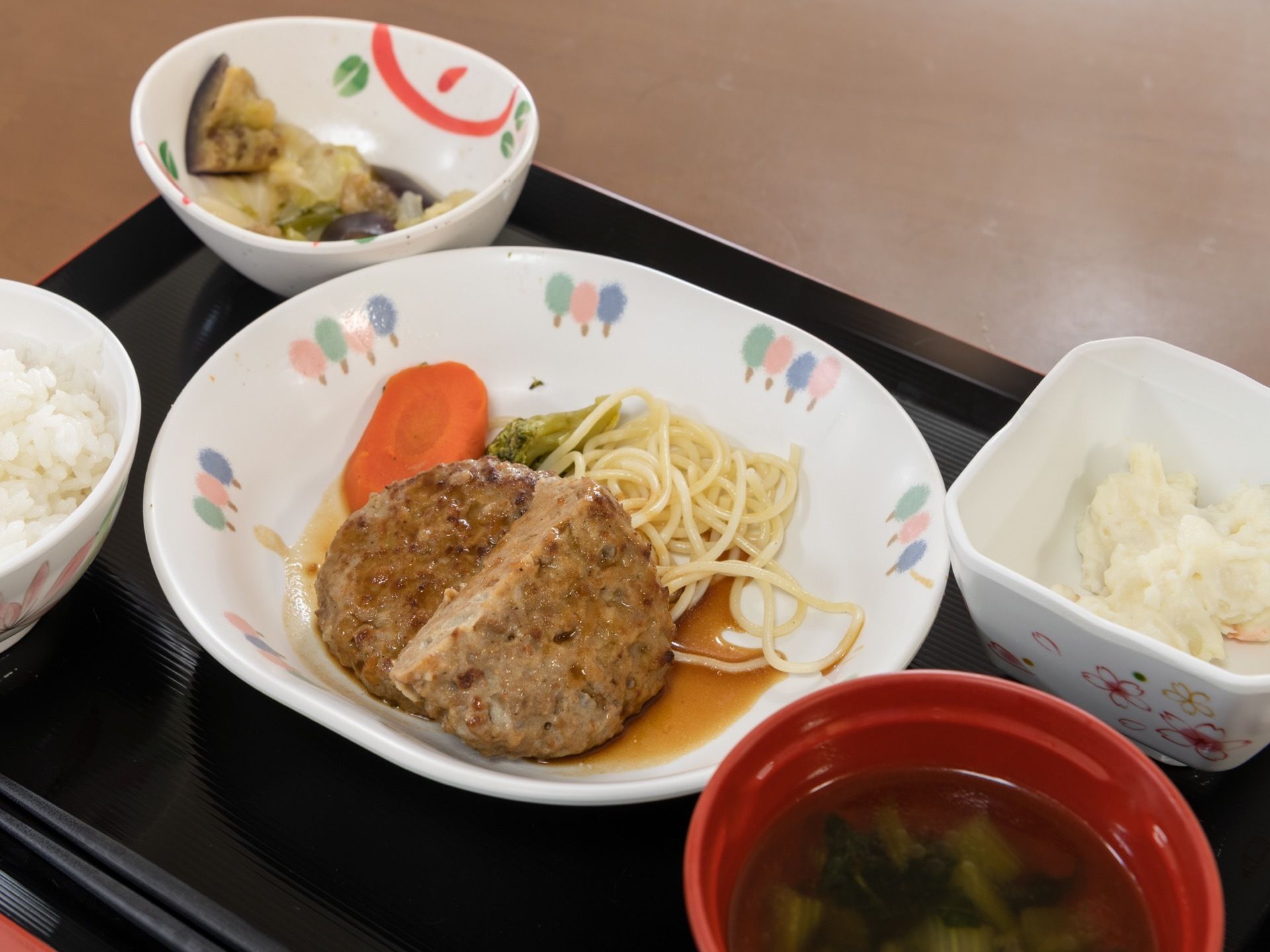 黒いお盆の上に、米飯にハンバーグ、ポテトサラダ、漬物が別のお皿に盛られて置いてある。味噌汁は、赤い器に入っている。