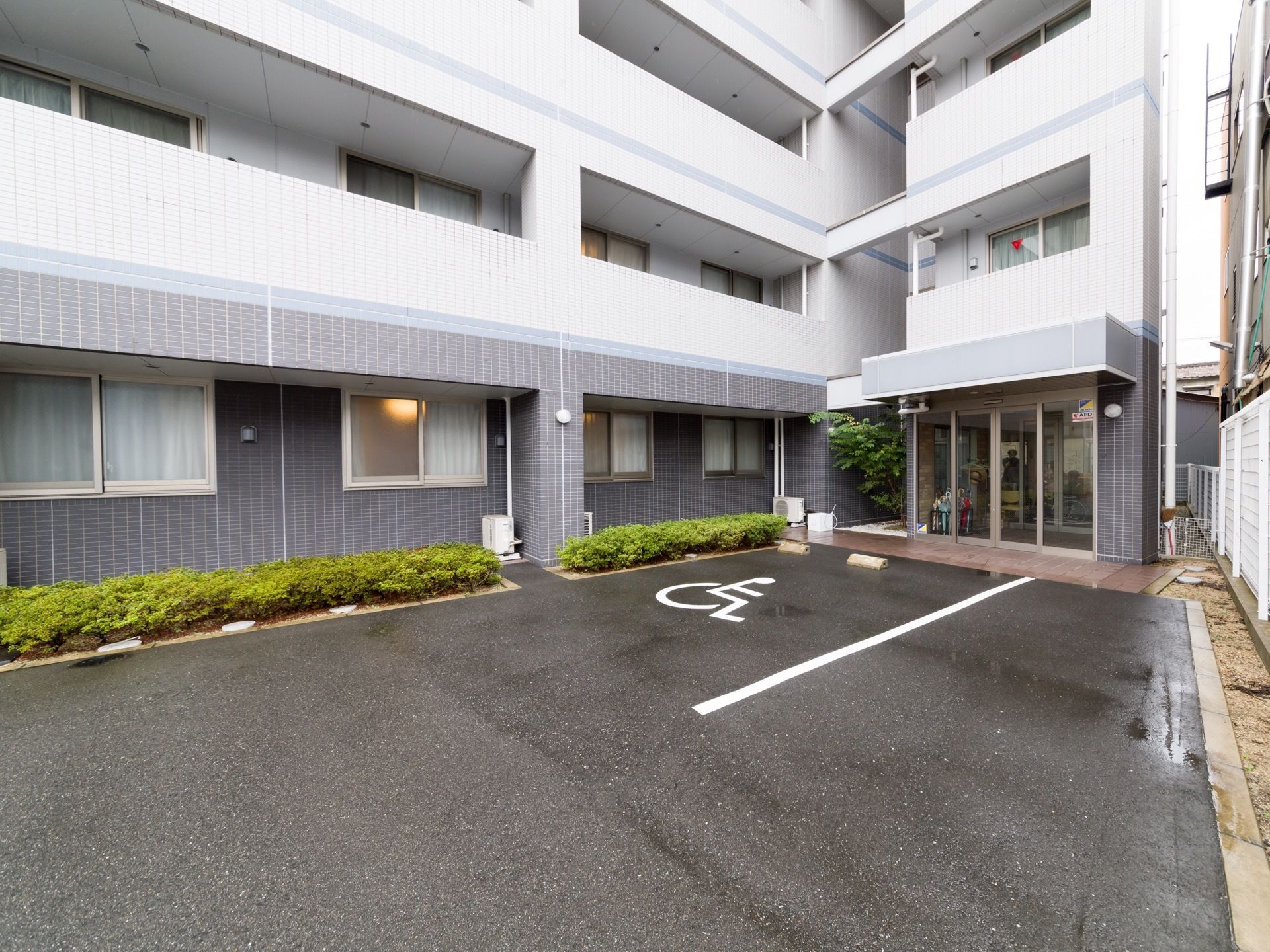 玄関前に車椅子専用の駐車スペースがある。玄関は２重構造の自動ドアになっている。建物の脇に植物が植えられている。