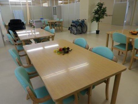 数台の淡い木目のダイニングテーブルと数脚の水色の椅子がゆとりをもって配置されている。２台の車椅子が壁際に並んで置いてある。