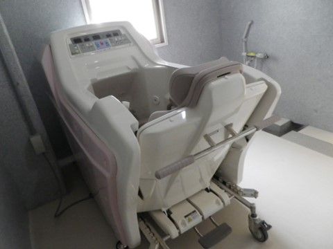 排水溝がついたベージュ色の床と、グレー色の壁に小窓がついた浴室である。キャスター付きの座椅子がついた介護浴槽器が置いてある。