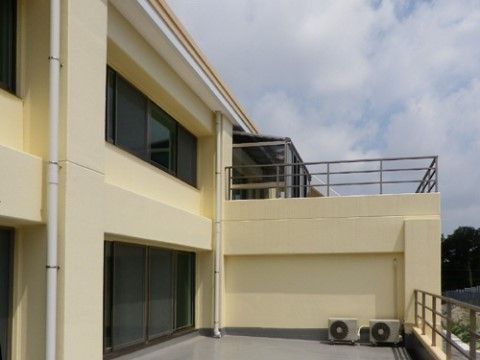 クリーム色の壁とグレー色の床の升目の大きな柵が取り付けられた空を臨める開放的な空間である。エアコンの室外機が２台置いてある。