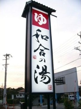 入浴施設の名称が入った大きな看板の向こう側に水色の柱の高床式の白い建物がある。電信柱から電線が伸びている。