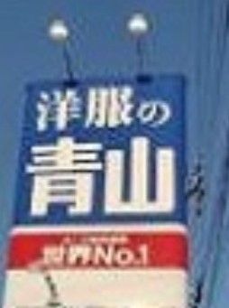 雲一つない青空のもとに、洋服店の名称が白文字で表示された２つの照明付きの大きな看板がある。看板の横に電線が見える。