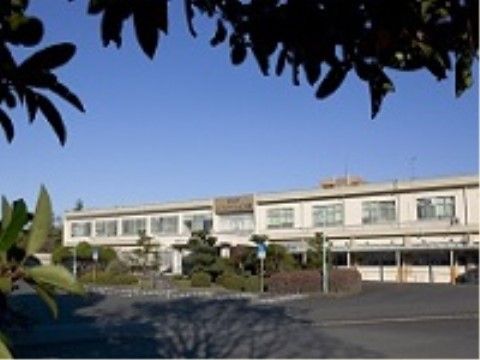 白い２階建ての建物の手前に舗装されたアスファルトの敷地があり、植栽スペースにたくさんの樹木が植えられている。