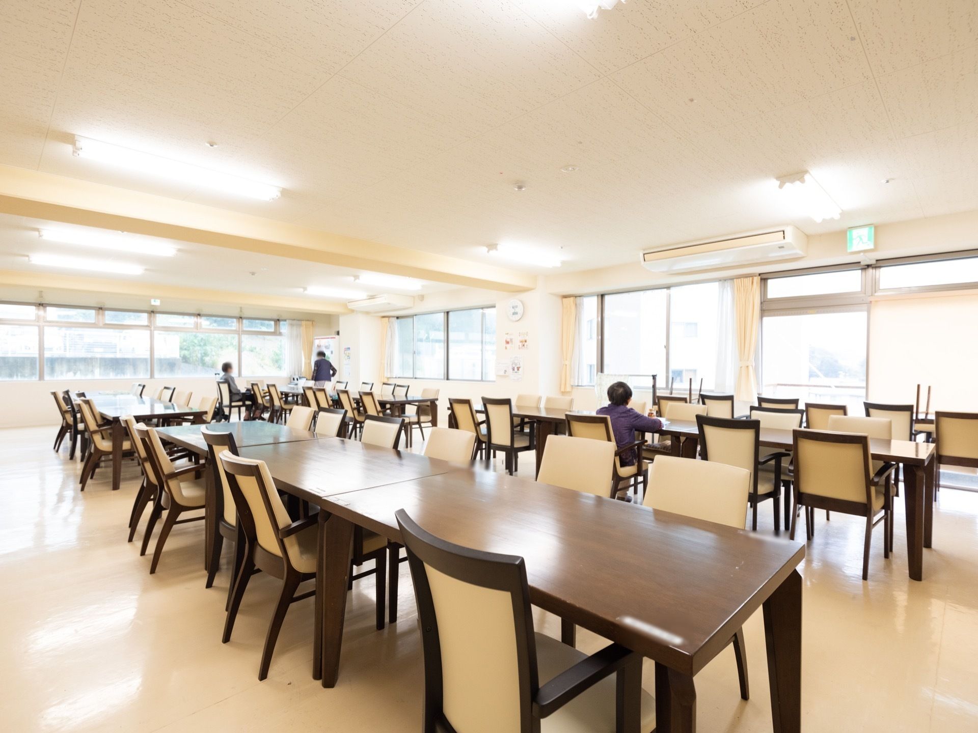 ２面の壁に多くの窓があってとても明るい食堂である。入居者様が２名、離れた位置にある椅子に座っている。