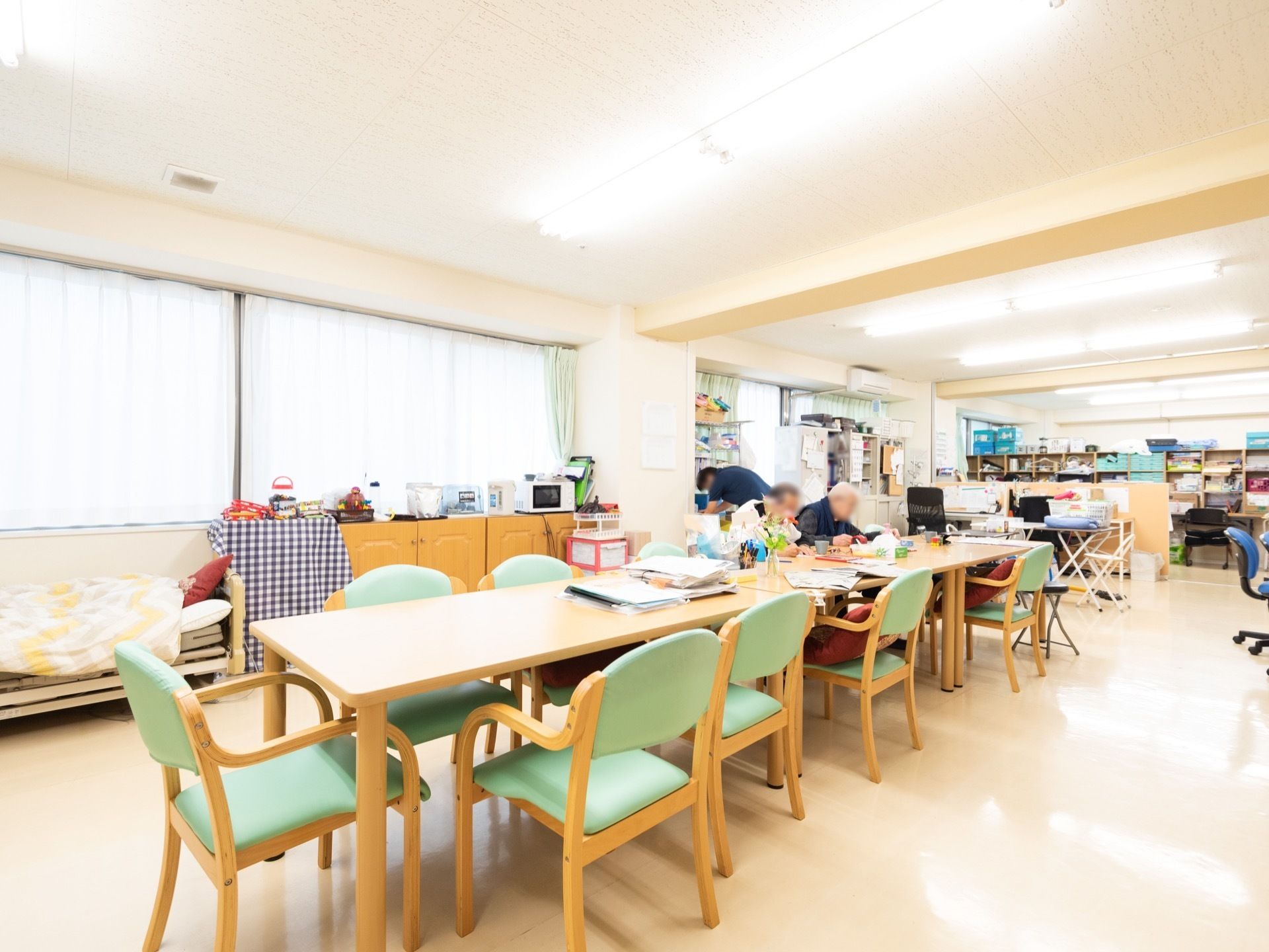 明るい色調のテーブルや椅子が置かれた共有スペースがある。男性の入居者様が２名、並んで座って机に向かっている。