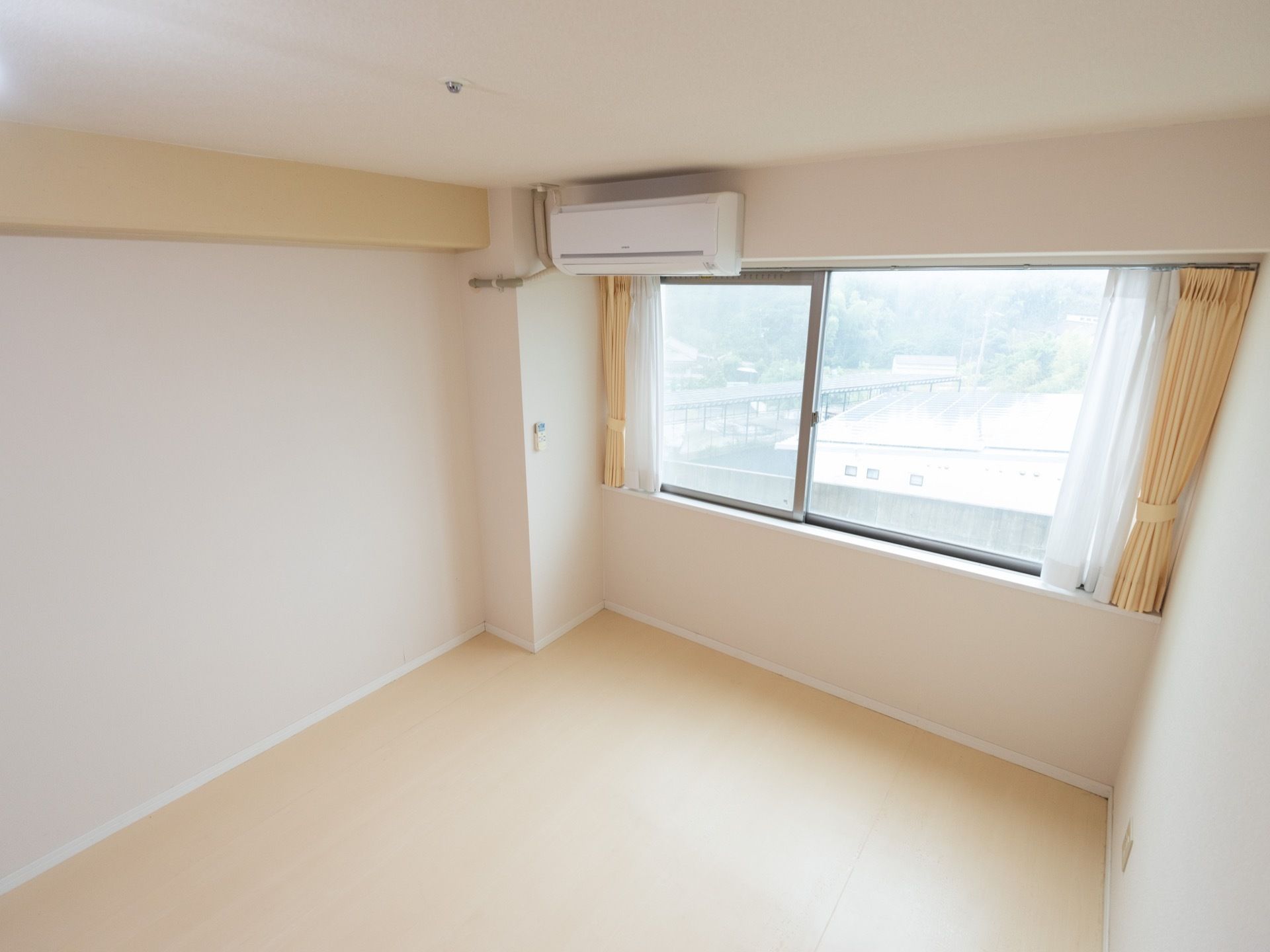 居室にある腰高の窓にレースと黄色のカーテンがかかっている。その上に白色のエアコンを設置。窓越しに近くの街並みや木々が見える。