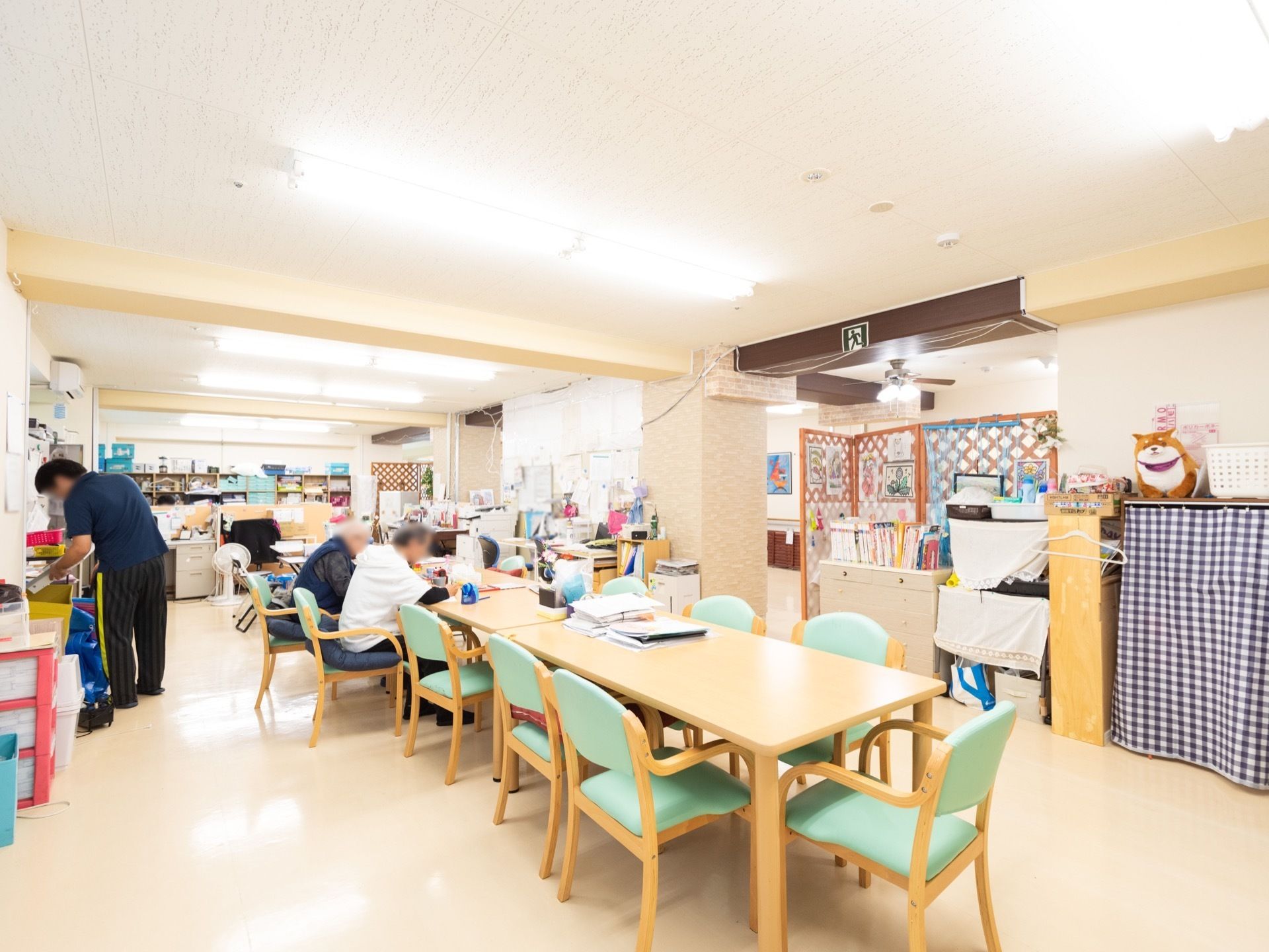 天井に多くの照明器具が設置された共有スペースがある。薄緑色の座面や背もたれがついた椅子に入居者様が座っている。