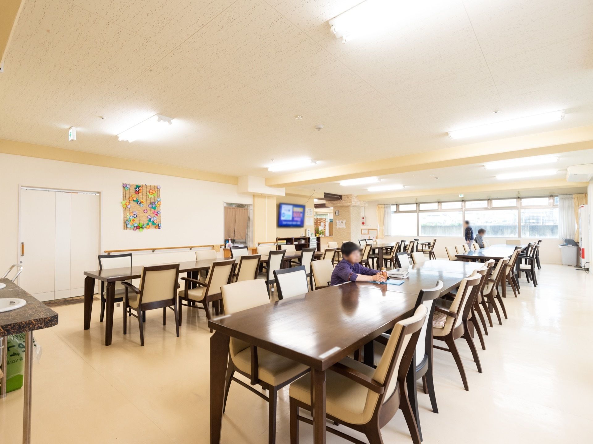 食堂の椅子に入居者様が２名座っている。１名の方が窓際を歩いている。壁に季節の花を紙で作った飾りが飾られている。