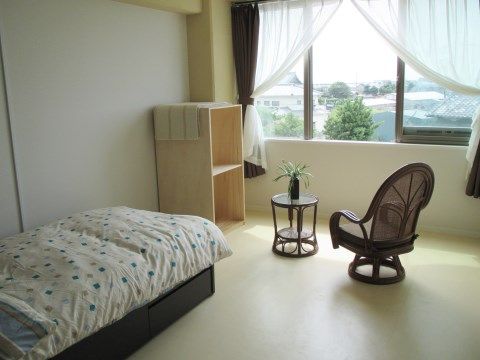 レースのカーテンがついた大きな窓のある居室。晴れた日は外の景色を楽しめる。ベッド、棚、机といすが備え付けてある。