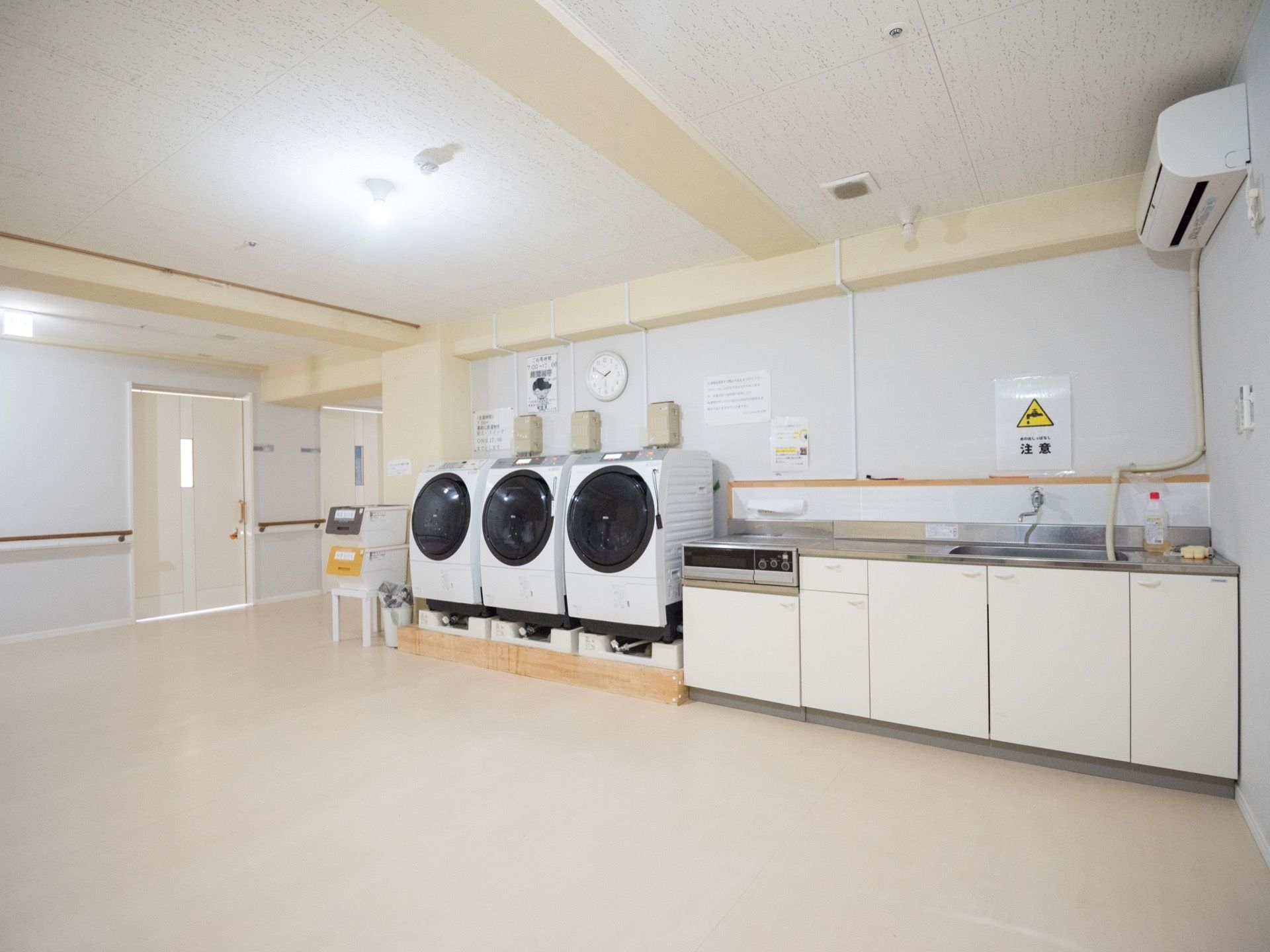 白い収納扉が取りつけられたシンクがあり、電気コンロが置いてある。黒い丸扉の洗濯機が３台並べられている。