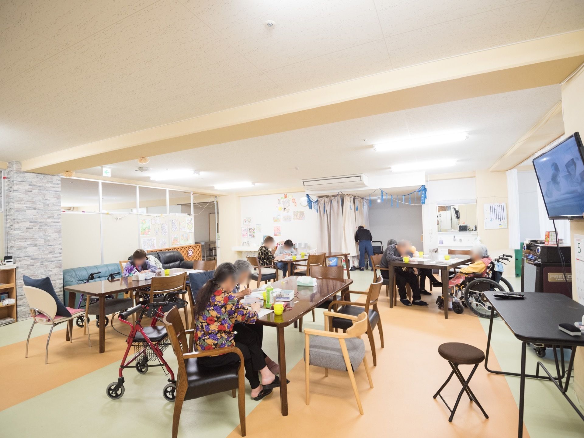 蛍光灯が点灯した広い部屋にたくさんのテーブルや椅子が置いてある。数名の入居者様が椅子や車椅子に腰を掛けている。