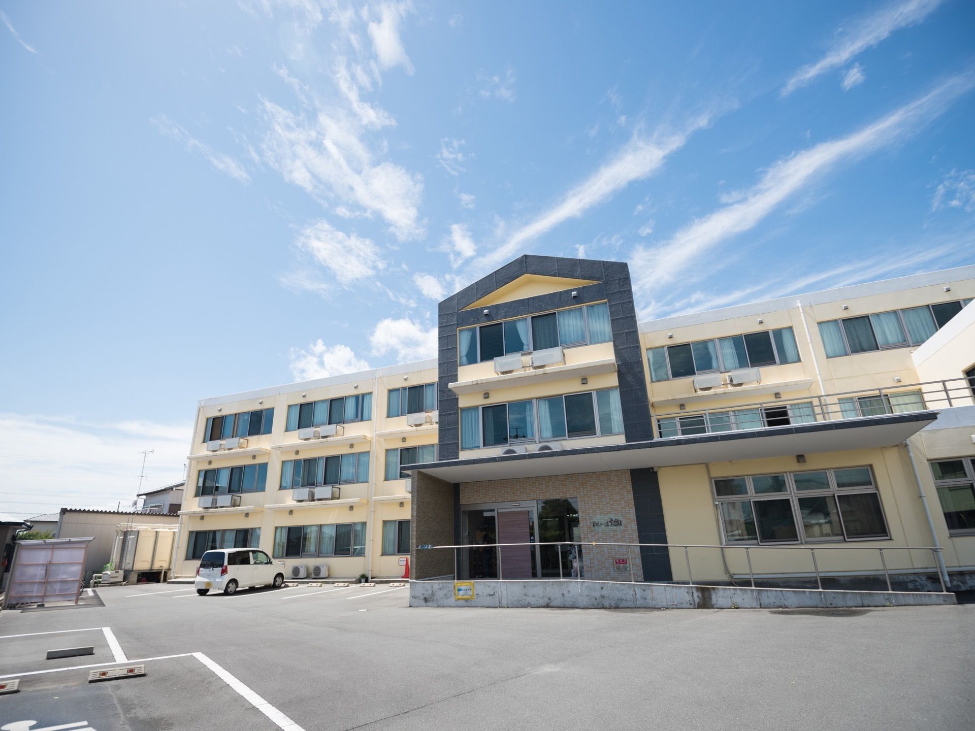 薄いクリーム色の３階建ての建物がある。建物の前は駐車スペースになっていて、白い軽自動車が１台停められている。