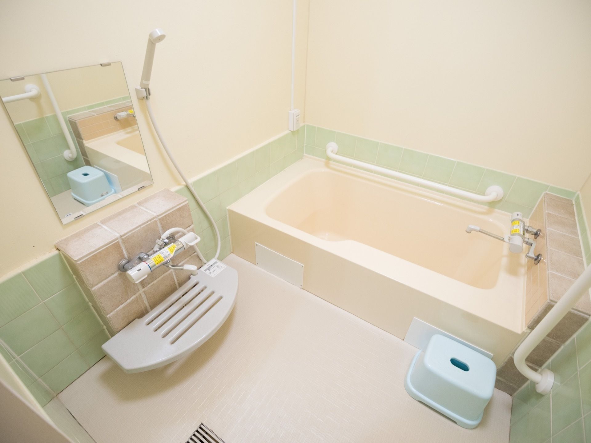 薄いピンク色の浴槽の隣の壁に白い手すりが設置されている。洗い場に水色の椅子があり、壁に鏡とシャワーが取りつけられている。