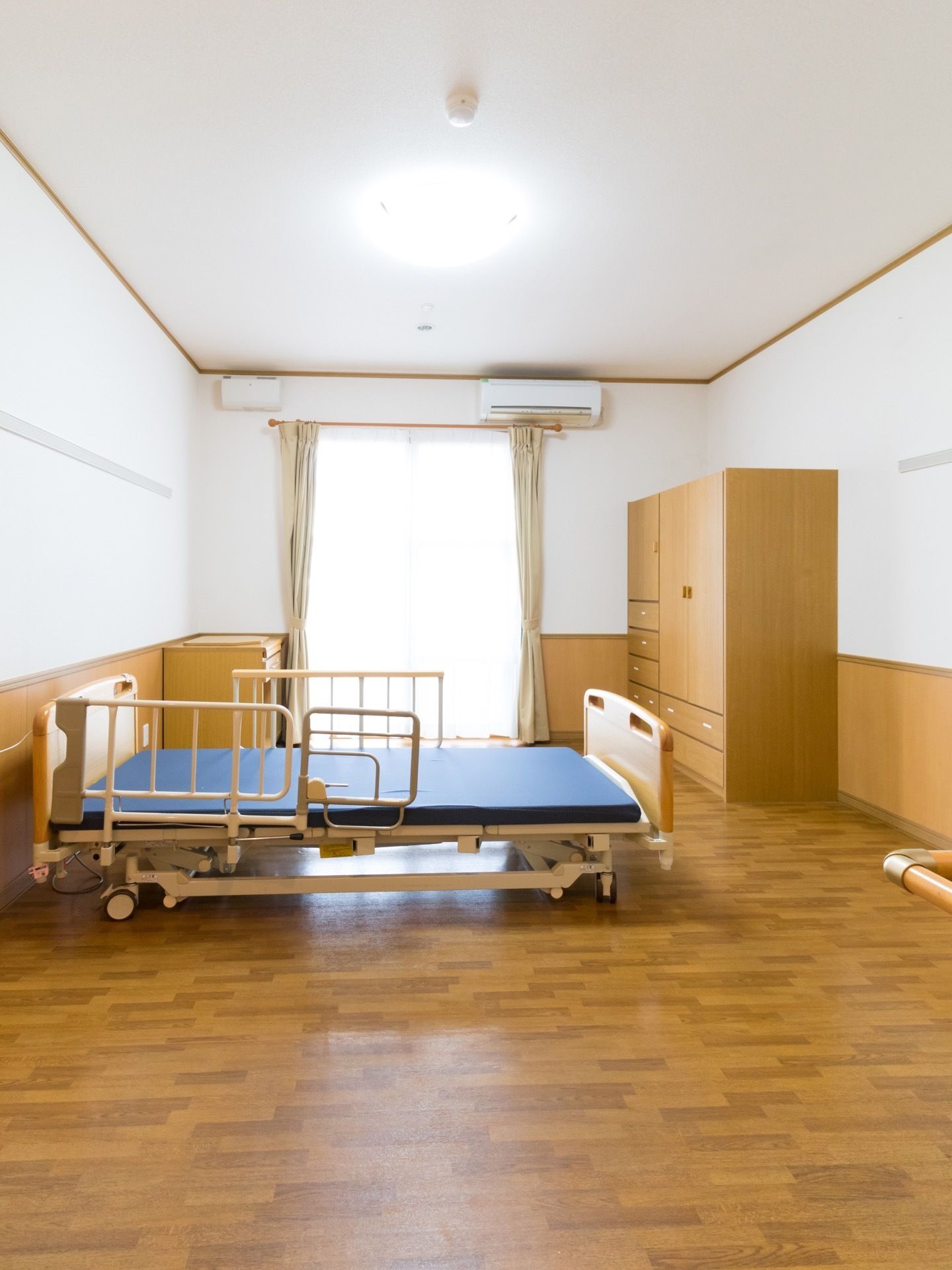 フローリング張りの室内に、チェストや介護ベッド、机が置かれている。窓の近くにエアコンが設置されている。