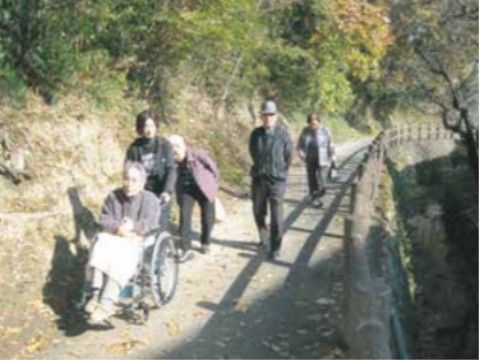 ４人の入居者様が山間の散歩道を歩いてこちらに向かっている。車椅子をご利用の方の後ろを女性スタッフが押している。