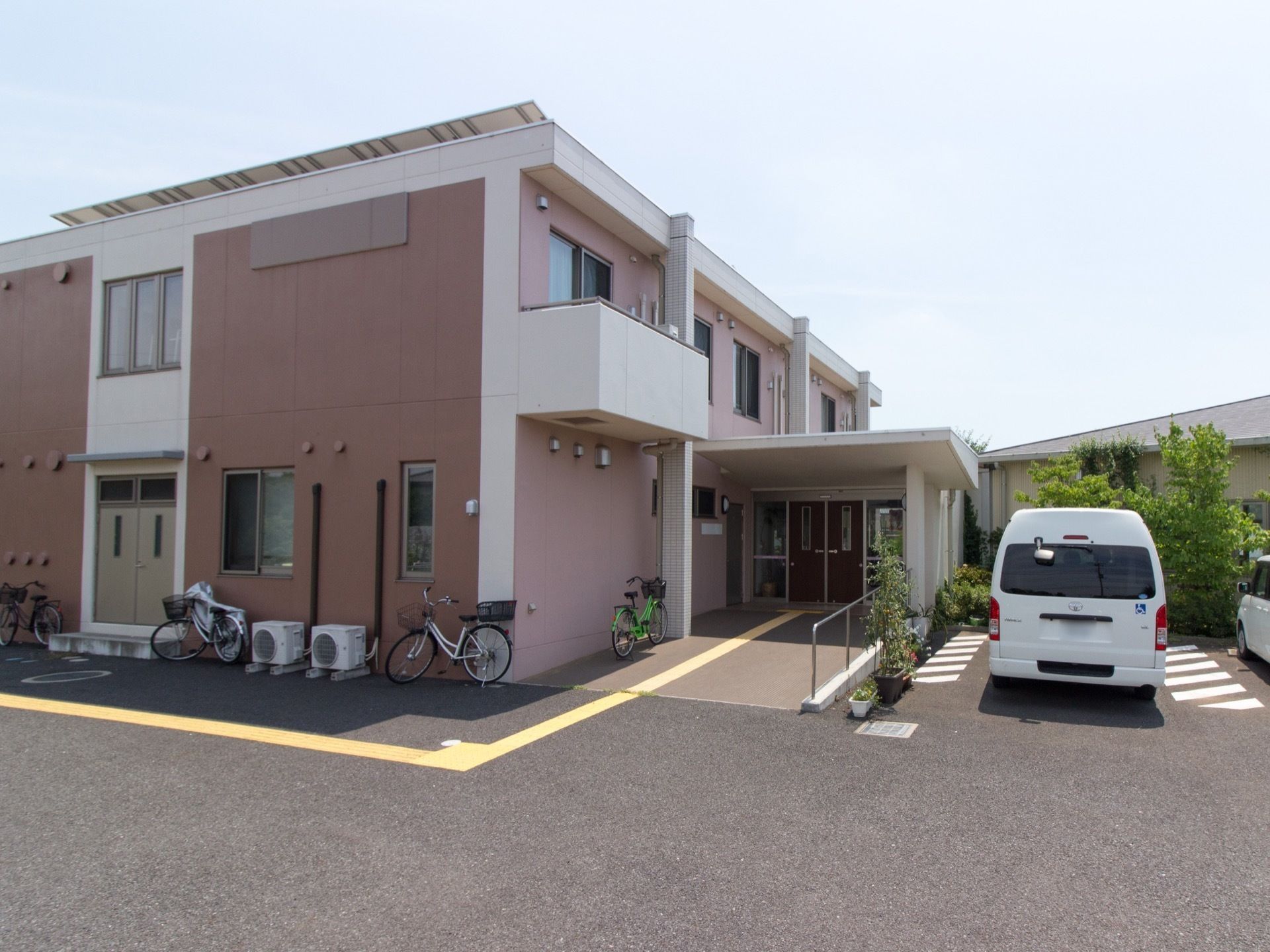 淡いピンク色の２階建ての建物である。玄関のひさしが突きだしている。玄関先まではスロープが設けられている。玄関横は駐車場となっている。