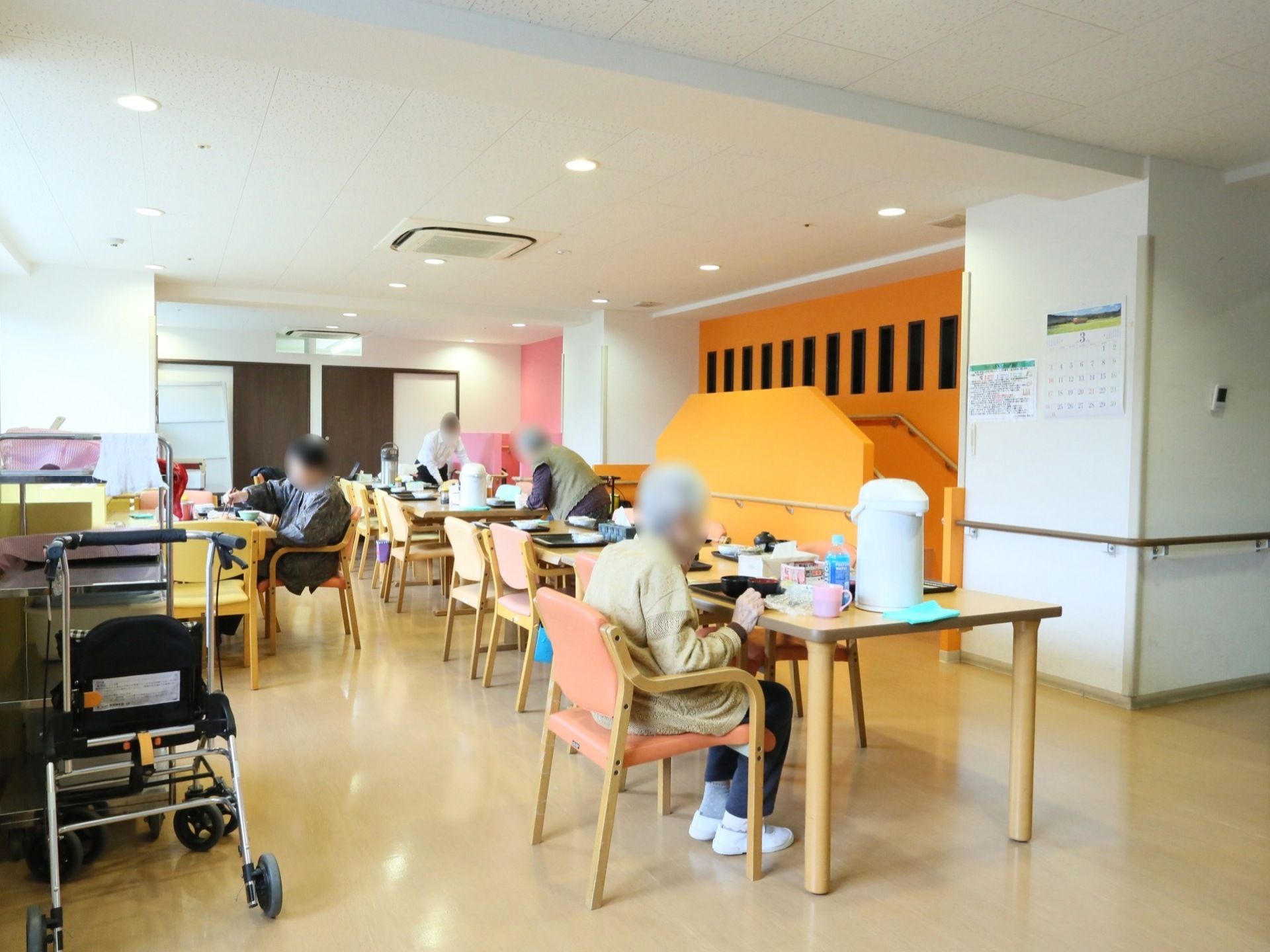 食堂に入居者様が集まって、食卓について食事を召し上がっている。奥の方では、スタッフが配膳作業を行っている。