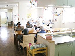 わっかの紙飾りが天井からぶら下がっていて、共有スペースでは入居者が思い思いの場所で過ごしている。手前にはキッチンがあり、ダイニングテーブルも置かれている。