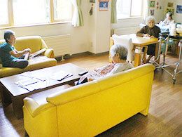 対面式に置かれた黄色いソファや奥のダイニングテーブルに入居者が座り思い思いに過ごしている。壁には入居者の作品が飾られ、館内はきれいに片付いている。