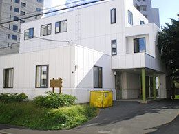 緑がたくさん周囲に植えられた白い3階建ての建物はか配ったデザイン。後ろの方には背の高いビルもある。玄関の横には黄色い箱が置かれている。
