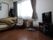 レースカーテンとカーテンが付いた大きな窓があるフローリングの部屋には、寝具付きのベッドやいすなどがある。