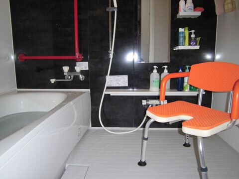 オレンジのシャワーチェアが置かれたモノトーンの浴室にはシャワーがあり、手すりも設置されている。
